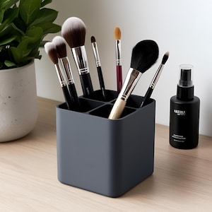 Könnte beinhalten: Ein grauer Make-up-Pinselhalter mit mehreren Fächern, gefüllt mit verschiedenen Make-up-Pinseln. Rechts befindet sich eine schwarze Sprühflasche mit weißem Text. Die Pinsel haben schwarze Griffe und weiße und braune Borsten. Der Halter steht auf einer hellen Holzoberfläche.