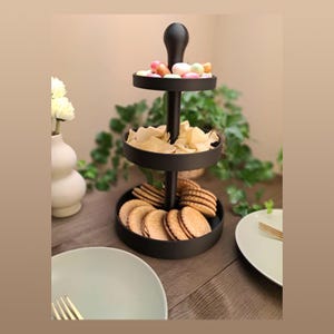 Op de afbeelding: Een zwart, drielaags serveerrek gevuld met snacks. De bovenste laag bevat kleurrijke snoepjes, de middelste laag chips en de onderste laag koekjes. De serveerstandaard staat op een houten tafel.