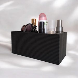 Op de afbeelding: Een zwarte, rechthoekige organizer met een geribbelde buitenkant, met diverse cosmetica-artikelen. Inhoud: zonnebril, lippenstift, deodorant en andere schoonheidsproducten. De organizer staat op een witte achtergrond.