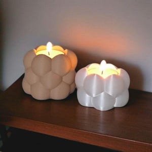 Op de afbeelding: Twee belvormige kandelaars met brandende kaarsen. Eén is beige, de andere wit. De houders staan op een donkerbruin houten oppervlak. De kaarsen geven een warme, uitnodigende gloed.