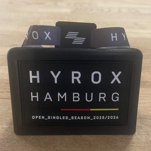 Può includere: Un badge rettangolare nero per eventi con cordino, che riporta il testo bianco HYROX HAMBURG, una linea orizzontale rossa e gialla e la scritta OPEN_SINGLES_SEASON_2025/2026 in basso. La parte superiore del badge mostra un piccolo logo bianco.
