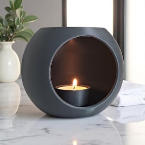 Op de afbeelding: Een donkergrijze, bolvormige kandelaar met een cirkelvormige opening. Een brandende waxinelichtkaars staat binnenin en geeft een warme gloed. De houder staat op een marmeren oppervlak, met een witte vaas en groen in de achtergrond.