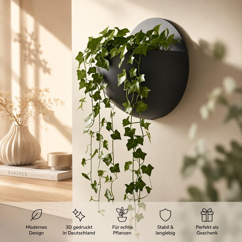 May include: A matte black, circular wall-mounted planter containing a trailing green ivy plant. The planter features a semi-circular opening at the top. Below the planter, text icons indicate features including 3D printed in Germany.