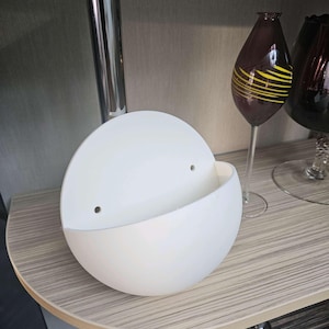 May include: A matte white, semi-spherical wall planter featuring a flat back panel with two small, circular mounting holes. The container has a curved, open-top design, providing a smooth, clean surface for holding small items or decorative plants.