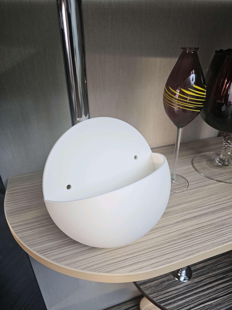 May include: A matte white, semi-spherical wall planter featuring a flat back panel with two small, circular mounting holes. The container has a curved, open-top design, providing a smooth, clean surface for holding small items or decorative plants.