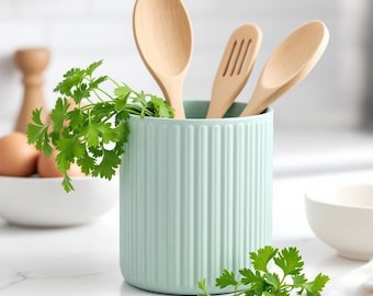 Può includere: Un portautensili in ceramica verde chiaro, a coste, con tre utensili da cucina in legno ed erbe verdi fresche. Il portautensili contiene un cucchiaio di legno, un cucchiaio scanalato e un altro cucchiaio di legno. La scena della cucina comprende una ciotola bianca e uova.
