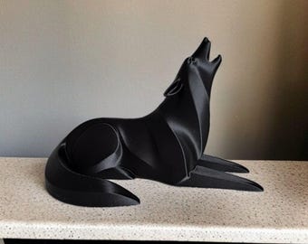 May include: A black sculpture of a howling wolf in a reclining position. The artwork has a modern, geometric design with smooth, curved lines. The wolf's head is raised, mouth open in a howl, and the fur appears sleek and polished.