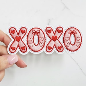 May include: Embroidered patch spelling "XOXO" in red and white. The letters are decorated with hearts and bows, perfect for Valentine's Day or expressing affection. The patch has a scalloped edge.