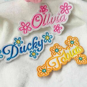 May include: Embroidered name patches in pink, blue, and yellow, each with a floral border. The names "Olivia", "Ducky", and "Tobias" are displayed in cursive script. The patches are on a white, textured surface.