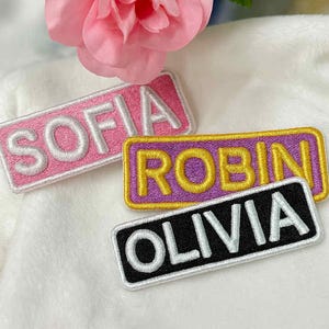 May include: Embroidered name patches in pink, purple, and black. The patches read "SOFIA", "ROBIN", and "OLIVIA" in white lettering. Each patch has a contrasting border. A pink rose is in the background.