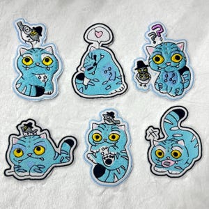 May include: Six embroidered patches featuring cartoon cats in various poses. The cats are light blue with yellow eyes and black accents. Each patch has a black border and a small bird or thought bubble. The patches are on a white surface.