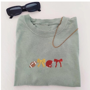 May include: Sage green sweatshirt with embroidered football, bow, helmet, and bow designs. Includes a gold chain necklace and black sunglasses.