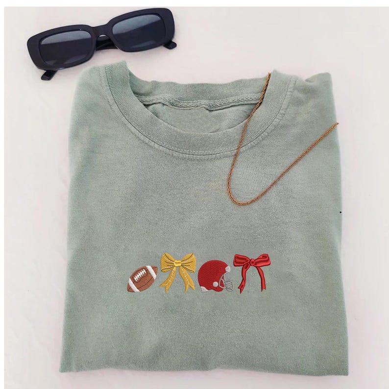 May include: Sage green sweatshirt with embroidered football, bow, helmet, and bow designs. Includes a gold chain necklace and black sunglasses.