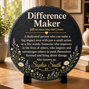 May include: A circular black slate plaque featuring the white text Difference Maker, a phonetic pronunciation, and a definition of the term. The design includes floral illustrations in white, yellow, and purple, with the name Sophia Smith printed below.