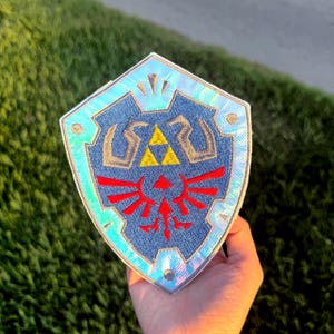 May include: A shield-shaped patch with a blue denim background and a silver iridescent border. The shield features a gold triangle and red emblem, reminiscent of a video game.