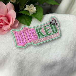 Op de afbeelding: Geborduurde patch met het woord "Wicked" in roze en groen, met een heksenhoed-icoon. De zin "For Good" staat onder het hoofdword. De patch heeft een lichtgroene rand en ligt op een witte, zachte ondergrond.