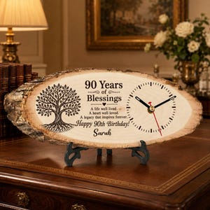 May include: A live edge wood slice clock featuring a printed tree illustration and the text 90 Years of Blessings, A life well lived, A heart well loved, A legacy that inspires forever, Happy 90th Birthday, and the name Sarah on a black stand.