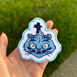 May include: Embroidered patch featuring a blue cat with a top hat. The cat has large eyes, whiskers, and a toothy grin. The patch is outlined in white and light blue, with a cloud-like shape.
