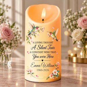 Puede incluir: Una vela de pilar LED cilíndrica en color crema suave con una llama parpadeante en la parte superior. La superficie muestra dos colibríes, detalles florales rosas y el texto: A LOVING THOUGHT A Silent Tear A CONSTANT WISH THAT You were Here Emma William 1960-2026.