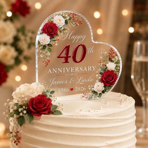 Può includere: Un topper per torta a forma di cuore in acrilico trasparente con il testo Happy 40th ANNIVERSARY James & Linda 1986 2026. Il topper è decorato con rose rosse e bianche, foglie verdi e piccole bacche, posizionato su una torta glassata bianca.