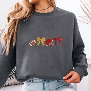 May include: A dark gray sweatshirt featuring embroidered football-themed designs. The designs include a football, a yellow bow, a red football helmet, and a red bow. The sweatshirt is a comfortable, casual style.