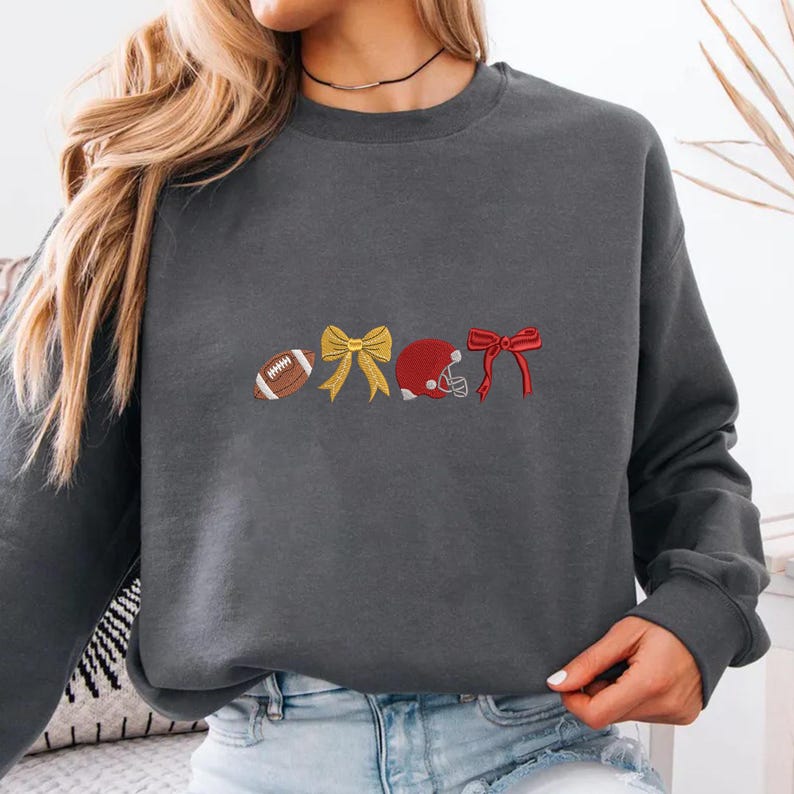 May include: A dark gray sweatshirt featuring embroidered football-themed designs. The designs include a football, a yellow bow, a red football helmet, and a red bow. The sweatshirt is a comfortable, casual style.