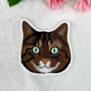 Puede incluir: Parche bordado con la cara de un gato. El gato tiene pelaje marrón con rayas más oscuras, ojos verdes y un hocico blanco. El parche está delineado con un borde blanco, ideal para ropa o accesorios.