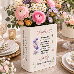 Pode incluir: Um vaso de cerâmica branco em forma de livro com o texto Chapter 50, um poema e o nome Emily em letra cursiva rosa. O vaso é decorado com ilustrações florais e pequenas borboletas, e está cheio com um buquê colorido de flores rosas e laranjas.