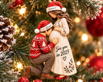 Può includere: Una decorazione natalizia da appendere che raffigura una persona con un cappello da Babbo Natale rosso con scritto Emma e una persona con un cappello rosso con scritto David che bacia un pancione. L'abito bianco riporta il testo See you in 2027.