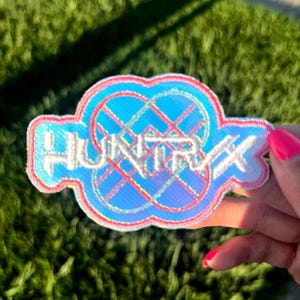 May include: A blue, red, and white embroidered patch with the word "HUNTRX" in white lettering. The patch has a unique shape with a light blue background and a red and white border. The patch is held against a green grassy background.