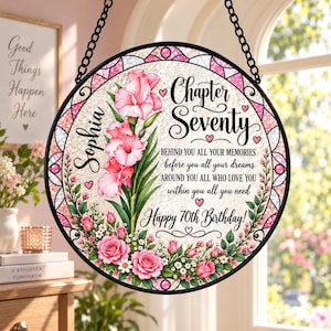 May include: A circular hanging sign with a black frame and chain, featuring a stained-glass style border in pink and white. The center displays pink gladiolus and roses, the name Sophia, and the text Chapter Seventy, Happy 70th Birthday.