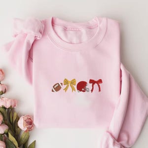 May include: A light pink sweatshirt with embroidered designs of a football, a gold bow, a red football helmet, and a red bow. The sweatshirt has long sleeves and a crew neck. The design is centered on the front of the shirt.