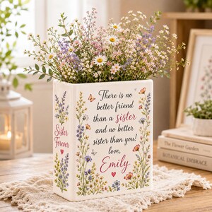 Puede incluir: Un jarrón rectangular de cerámica blanca lleno de flores silvestres. El frente presenta el texto: There is no better friend than a sister and no better sister than you! love, Emily. Los lados están decorados con motivos florales y el texto Sister Forever.