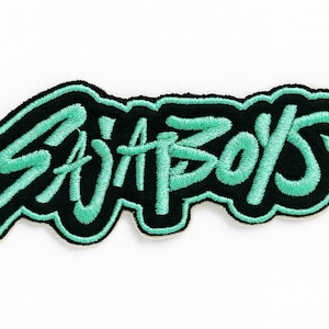 May include: Embroidered patch with the text "SADA BOYS" in a stylized font. The lettering is a bright teal color, outlined in black, and set against a white background. The patch has a black border.