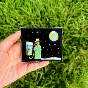 May include: Embroidered patch featuring a cartoon scene of a person in a green coat and hat, holding a phone, with a moon and stars in the night sky. The patch is rectangular with a black background and white border.