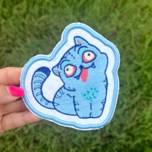Puede incluir: Un parche bordado azul con un gato de dibujos animados con ojos rojos y una lengua rosa. El gato está delineado en blanco y azul claro. El parche se sostiene sobre un fondo de hierba verde.