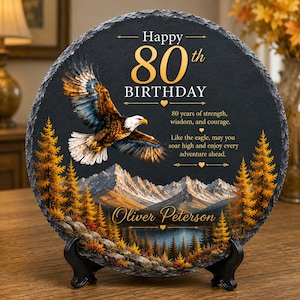 May include: A round black slate plaque featuring a bald eagle in flight over a mountain landscape with golden trees. The text reads Happy 80th BIRTHDAY, 80 years of strength, wisdom, and courage. Like the eagle, may you soar high and enjoy every adventure ahead. Oliver Peterson.