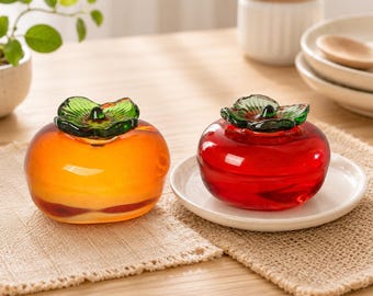 May include: Two glass trinket boxes shaped like fruits, one orange and one red, each featuring a translucent body and a detailed green glass leaf lid. These decorative containers are smooth, polished, and designed with a rounded, bulbous form.