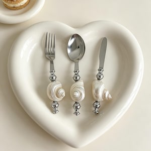 May include: A stainless steel fork, spoon, and knife set with seashell handles, arranged on a heart-shaped white plate. The silverware has a decorative silver accent between the shell and the utensil head. The shells are white and pearlescent.