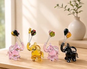 May include: Four glass elephant figurines in pink, yellow, pink, and black, each holding thin metal wires topped with small, colorful glass balloon-shaped spheres. The elephants are standing in a row, showcasing translucent bodies and trunks.