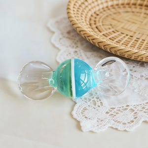 May include: A clear glass candy-shaped ornament with a blue and white stripe in the center. The ornament has rounded ends and is resting on a white lace doily. A woven basket is in the background.