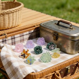 Pode incluir: Uma coleção de presilhas de cabelo em forma de flor em várias cores, incluindo rosa, roxo, verde e amarelo. As presilhas estão dispostas sobre um tecido xadrez dentro de uma cesta de piquenique, com uma caixa de metal e uma cesta de vime no fundo.