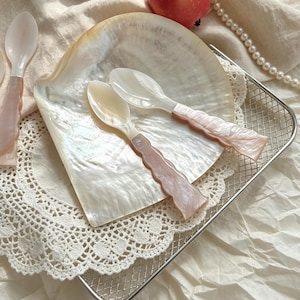 Puede incluir: Dos cucharas decorativas con cuencos nacarados y mangos rosas descansan sobre una gran concha iridiscente. Las cucharas están colocadas sobre un mantelito de encaje blanco dentro de una pequeña cesta de alambre plateado. Un collar de perlas y una manzana roja están en el fondo.