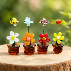 May include: Four miniature glass ornaments featuring brown pots with flowers and small creatures on thin metal stems. The figures include a yellow bee, a light blue insect, a purple bird, and a red bird, each perched above colorful glass petals.