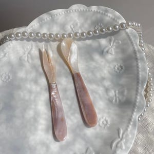 Puede incluir: Un juego de servir decorativo con tenedor y cuchara con mangos nacarados rosas y blancos. Los utensilios descansan sobre un plato blanco en relieve, con un collar de perlas en el fondo. Ideal para servir aperitivos.