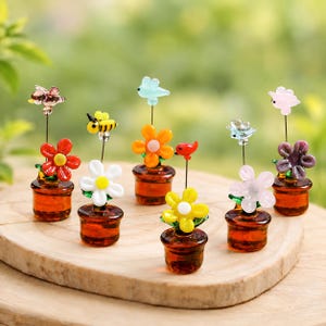 May include: A collection of six miniature glass flower pots, each featuring a small amber-colored base and a thin metal stem topped with a tiny glass figure, including various colorful flowers, a yellow and black bee, and small birds.