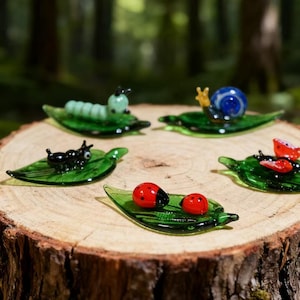 May include: A collection of miniature glass figurines of insects, including a caterpillar, snail, ant, and ladybugs, each resting on a green glass leaf. The ladybugs are red with black spots. The figurines are displayed on a wooden surface.