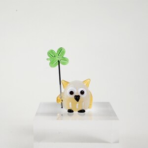 May include: A small, glass owl figurine with a white body, yellow accents, and large black eyes. It holds a green four-leaf clover on a black stem. The owl is displayed on a clear, square block.