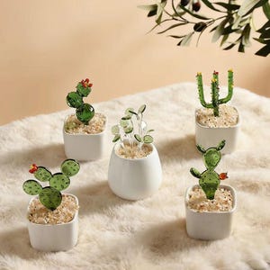 May include: Five decorative glass cactus plants in white square and oval pots. The glass cacti are green with red and yellow accents. The pots are filled with small, light-coloured pebbles. The plants are arranged on a soft, white surface.