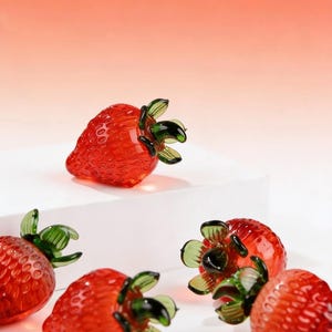 May include: A collection of glass strawberry ornaments, each with a red body and green leaves. The strawberries have a textured surface. The background is a gradient of peach and white. The ornaments are displayed on a white surface.
